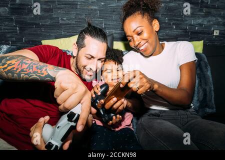 Fröhliche Mutter mit Smartphone, während Vater und Tochter spielen Videospiel auf dem Sofa zu Hause