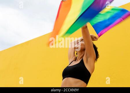 Glückliche Frau winkt Regenbogenfahnen, während sie gegen die gelbe Wand steht In der Stadt Stockfoto