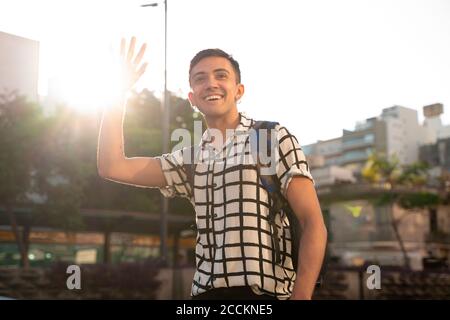 Lächelnder junger schwuler Mann winkt, während er in der Stadt gegen steht Himmel Stockfoto