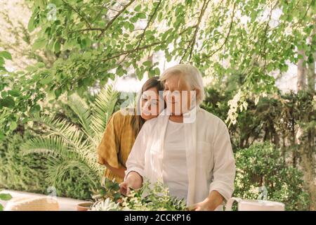 Tochter umarmt ältere Mutter steht gegen Pflanzen und Baum in Hof Stockfoto