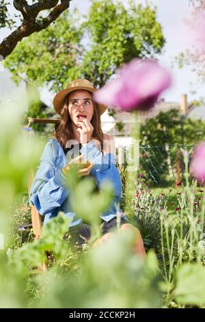 Junge Frau trägt Hut essen Erdbeere, während im Gemüse sitzen Garten Stockfoto