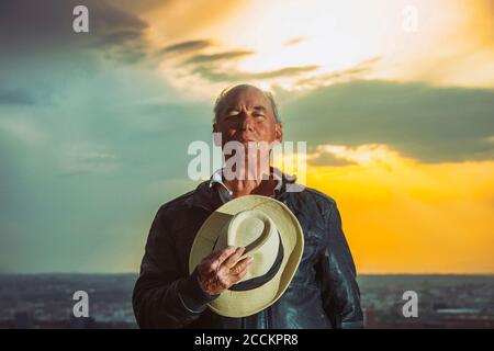 Älterer Mann hält Hut gegen bewölkten Himmel während des Sonnenuntergangs Stockfoto