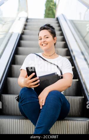 Porträt einer lächelnden kurvigen jungen Frau, die auf der Rolltreppe sitzt Mit Mobiltelefon Stockfoto