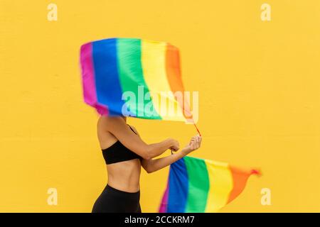 Frau winkt Regenbogenfahnen, während sie gegen die gelbe Wand steht Stadt Stockfoto