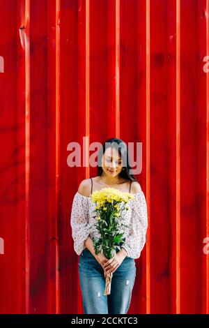 Junge Frau hält Blumen, während sie gegen die rote Wand steht Stockfoto