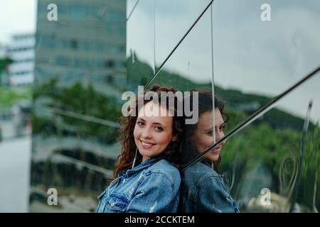 Lächelnde junge Frau, die sich an die Spiegelwand lehnt Stockfoto