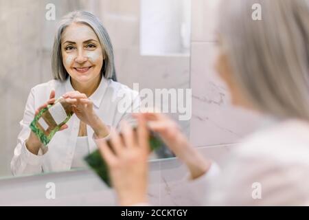 Rückansicht, Reflexion im Spiegel der attraktiven Senior grauhaarige Frau Anwendung feuchtigkeitsspendende Anti-Falten-Gesichtsmaske auf ihr Gesicht, während sie im Stehen Stockfoto