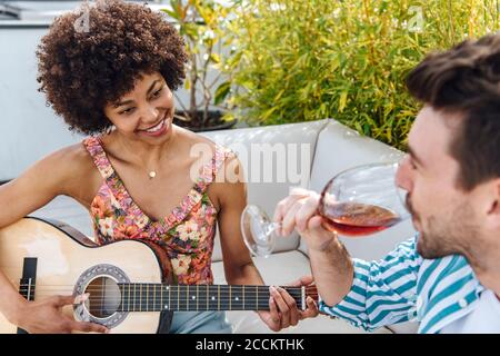 Glückliche Frau spielt Gitarre für Mann trinken Rotwein an Penthouse-Terrasse Stockfoto