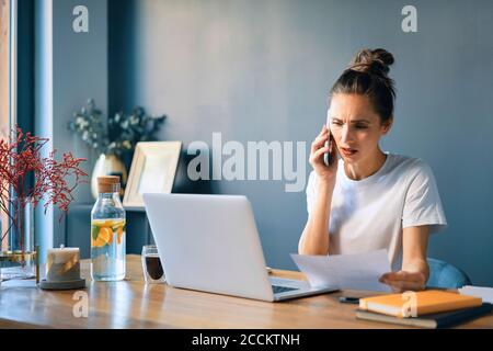 Junge Frau hält Dokument reden über Smartphone am Schreibtisch Im Heimbüro Stockfoto