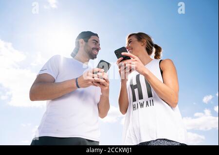 Lächelndes Paar hält Smartphones, während sie einander anguckend Stehen gegen den Himmel an sonnigen Tagen Stockfoto