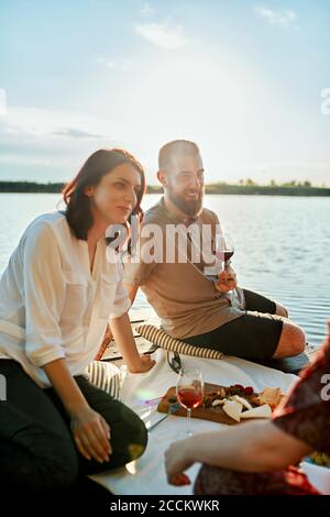 Freunde, die bei Sonnenuntergang am Steg an einem See picknicken Stockfoto