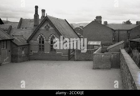 1950er Jahre, historisches Bild aus dieser Zeit zeigt die Dorfschule, Gebäude und geschlossenen Spielplatz, Llandyssil, Powys, Wales. Die viktorianische Schule wurde 1866 gebaut, entworfen von Thomas Henry Wyatt, einem anglo-irischen Architekten, der eine herausragende Karriere hatte, wurde 1870 zum Präsidenten des Royal Institute of Briitsh Architects gewählt und erhielt 1873 die Royal Gold Medal for Architecture. Die Schule schloss 1950 und Teile wurden erst 2010 durch eine Spendenaktion gerettet, um die Schulhalle zu einem lokalen Gemeindezentrum zu machen. Stockfoto