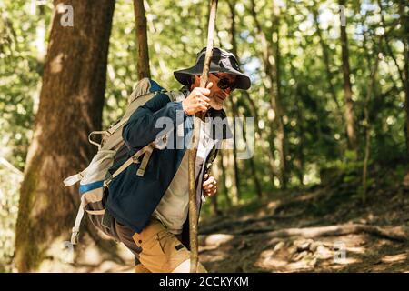 Seitenansicht eines reifen Mannes mit Rucksack, der auf einem Pfad durch Bäume läuft. Männlicher Rucksacktourist, der mit einem Stock durch Bäume wandert. Stockfoto