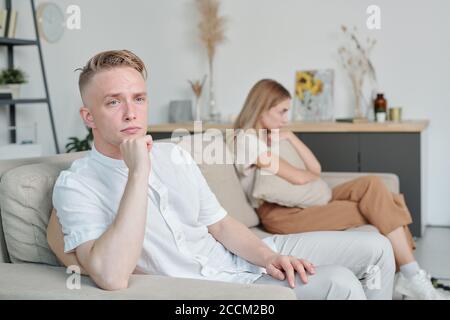 Beleidigter junger Mann, der mit seiner unglücklichen Frau auf der Couch sitzt Im Hintergrund Stockfoto