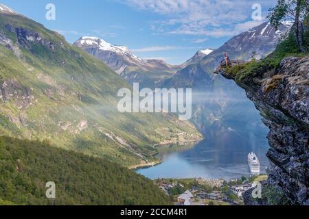 GEIRANGER, NORWEGEN - 2016. JUNI 12. Mann, der am Aussichtspunkt Flydalsjuvet am atemberaubenden UNESCO Geiranger Fjord sitzt, Stockfoto