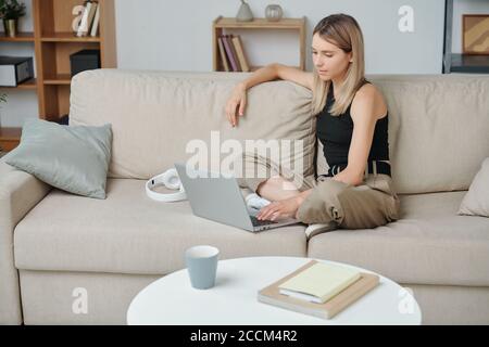 Zeitgenössische junge blonde Frau mit gekreuzten Beinen mit Laptop an Zu Hause Stockfoto
