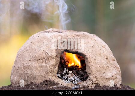 Ein kleiner, primitiver Ofen aus Schlamm. Ein Feuer brennt innen und Rauch steigt aus einem Loch in der Spitze. Stockfoto