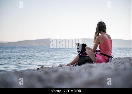 Junge Frau sitzt am schönen Kiesstrand mit ihrem schwarzen Schäferhund neben ihr liegen. Stockfoto