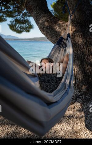 Junge Frau liegt und ruht in einer Hängematte unter einer Kiefer am Meer. Stockfoto
