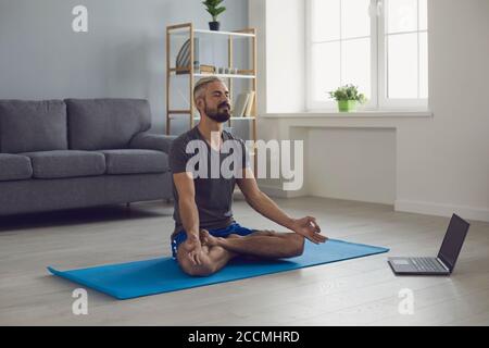 Online Yoga-Kurs. Friedlicher junger Mann, der morgens vor dem Laptop zu Hause Meditationspraxis macht. Life Balance Konzept Stockfoto