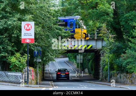 Northern Rail Personenverkehr Zug, Reisen vorbei Bahnhof Zeichen auf niedrige Eisenbahnbrücke & Auto auf ländlichen Dorf Straße - Burley, Yorkshire, England, Großbritannien Stockfoto