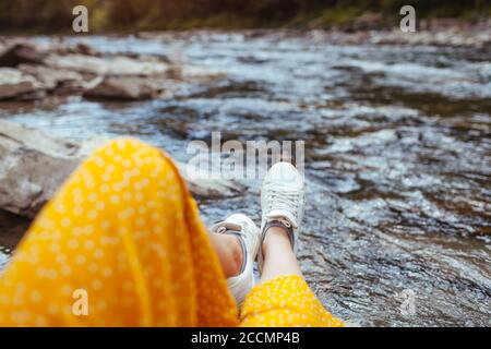 Frau entspannt am Bergfluss und genießt die Landschaft. Reisender sitzt auf Felsen. Sommerurlaub. Nahaufnahme der Schuhe Stockfoto