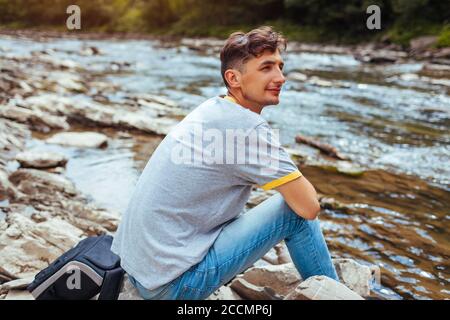 Tourist Mann entspannen am Bergfluss genießen natürliche Landschaft. Reisender Rucksacktourist sitzt auf Felsen. Sommerurlaub Stockfoto