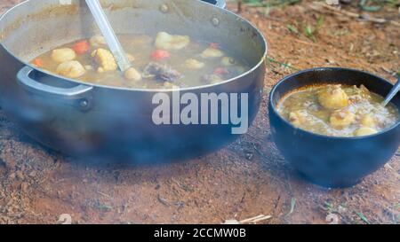 Ein Huhn und Dumplin Bouillion oder Suppe auf einem gekocht Holzfeuer im Freien in einem Metalltopf und Eine schwarze Keramikschale, die auf dem Schmutz ruht Stockfoto