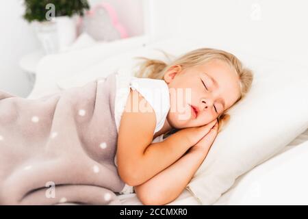 Kleines blondes Mädchen schläft auf weißem Bett mit den Händen unter ihrer Wange. Unbeschwert, Kindheit. Stockfoto