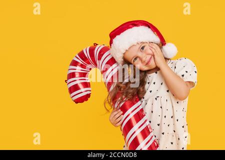 Ein niedliches kleines Mädchen Kind in einem eleganten Kleid und einem Weihnachtsmann Hut hält eine aufblasbare Form eines Zuckerrohrs auf einem gelben Hintergrund. 2021 Neujahr con Stockfoto
