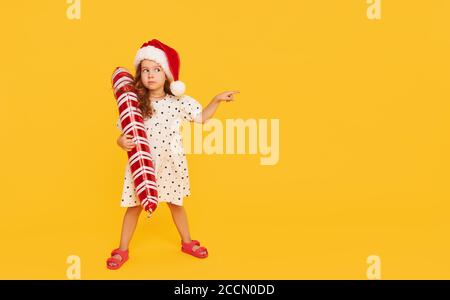 Ein niedliches kleines Mädchen Kind in einem eleganten Kleid und einem Weihnachtsmann Hut mit aufblasbarer Form eines Zuckerrohrs auf einem gelben Hintergrund. Zeigt die Hand nach links. 202 Stockfoto