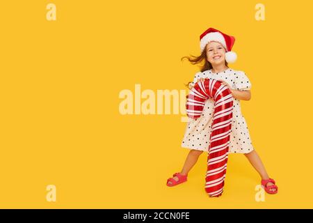 Ein niedliches kleines Mädchen Kind in einem eleganten Kleid und einem Weihnachtsmann Hut hält eine aufblasbare Form eines Zuckerrohrs auf einem gelben Hintergrund. 2021 Neujahr con Stockfoto