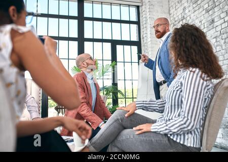 Selbstbewusster Geschäftsmann, der auf Flipcharts schreibt, präsentiert das neue Projekt in der Vorstandsetage bei der Unternehmenssitzung. Serious Mentor Auditor spricht mit Partnern Stockfoto