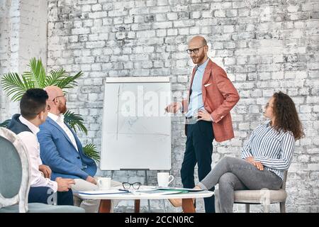 Selbstbewusster Geschäftsmann, der auf Flipcharts schreibt, präsentiert das neue Projekt in der Vorstandsetage bei der Unternehmenssitzung. Serious Mentor Auditor spricht mit Partnern Stockfoto