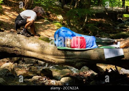 Paar Wanderer entspannen auf gefallenen Baumstamm über Berg Fluss Bach im sonnigen Sommer als shinrin Yoku Wald Baden und Natur Eskapismus. Stockfoto