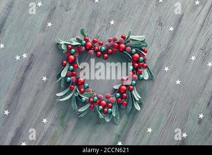 Dekorativer Xmas Mistelkranz mit roten Beeren und grünen Blättern in Herzform. Hölzerne Weihnachten Hintergrund mit Papier Stern Konfetti verziert. Stockfoto