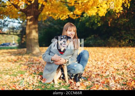 Fröhliche junge kaukasische Frau, die auf dem Boden sitzt und den Hund umarmt. Besitzer, der mit Haustier am Herbstfalltag spazieren geht. Beste Freunde, die im Freien Spaß haben. Freundschaft von h Stockfoto