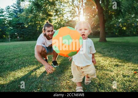 Junger Vater spielt Ball mit Kleinkind Baby Junge im Freien. Elternteil verbringt Zeit zusammen mit Kind Sohn im Park. Authentischer Lifestyle zarter Moment. Stockfoto