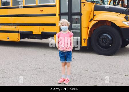 Glücklich kaukasischen Mädchen Student trägt Gesichtsmaske in der Nähe von gelben Bus. Kind mit persönlicher Schutzausrüstung im Gesicht. Bildung und zurück zur Schule Stockfoto