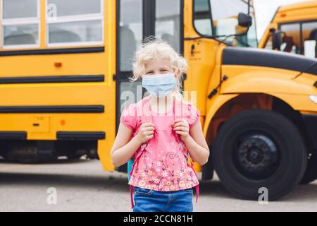 Glücklich kaukasischen Mädchen Student trägt Gesichtsmaske in der Nähe von gelben Bus. Kind mit persönlicher Schutzausrüstung im Gesicht. Bildung und zurück zur Schule Stockfoto