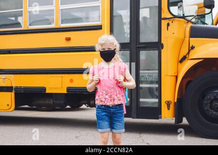 Glücklich kaukasischen Mädchen Student trägt Gesichtsmaske in der Nähe von gelben Bus. Kind mit persönlicher Schutzausrüstung im Gesicht. Bildung und zurück zur Schule Stockfoto