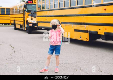Glücklich kaukasischen Mädchen Student trägt Gesichtsmaske in der Nähe von gelben Bus. Kind mit persönlicher Schutzausrüstung im Gesicht. Bildung und zurück zur Schule Stockfoto