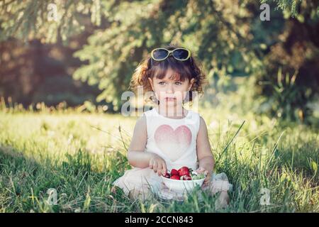 Nette liebenswert kaukasischen Kleinkind Baby Mädchen auf Gras sitzen und essen rote Beeren Früchte. Lustige Kind mit Sonnenbrille im Park mit gesunden Snack Stockfoto