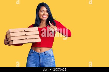 Hispanische Frau mit langen Haaren halten Lieferung Pizza Box lächelnd glücklich und positiv, Daumen nach oben tun ausgezeichnet und Genehmigung Zeichen Stockfoto