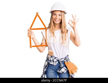 Schöne kaukasische Frau mit blonden Haaren trägt Architekt Hardhut und hält Werkzeuge tun ok Zeichen mit Fingern, lächelnd freundlich Gestik excellen Stockfoto