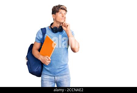 Junger gutaussehender Mann trägt Student Rucksack und Kopfhörer halten Buch ernstes Gesicht Denken über Frage mit Hand auf Kinn, nachdenklich über confu Stockfoto