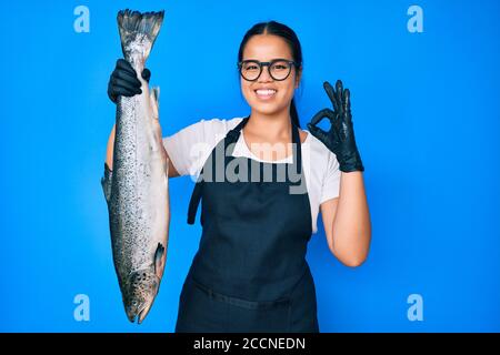 Junge schöne asiatische Mädchen Fischhändler Verkauf frischer roher Lachs tun ok Zeichen mit Fingern, lächelnd freundlich gestikulieren ausgezeichnete Symbol Stockfoto