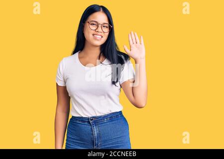 Junge schöne asiatische Mädchen tragen lässige Kleidung und Brille waiving sagen hallo glücklich und lächelnd, freundliche Willkommensgeste Stockfoto