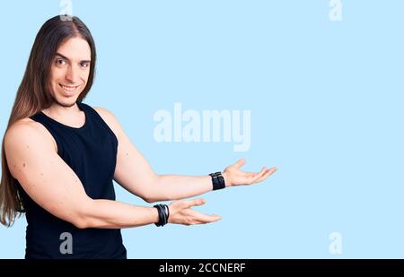 Junge Erwachsene Mann mit langen Haaren tragen Goth Stil mit Schwarze Kleidung, die zum Eintreten einlädt, lächelt natürlich mit offener Hand Stockfoto