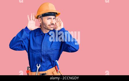 Junge hispanic Mann trägt Arbeiteruniform versucht, beide Hände auf Ohr Geste hören, neugierig auf Klatsch. Hörproblem, taub Stockfoto